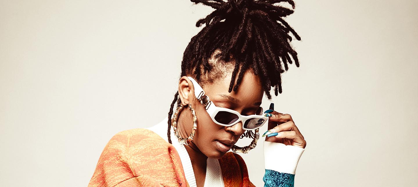 The SheJay poses for a photo. She is wearing white sunglasses and large gold hoops. Her hair is braided and tied high on her head. She looks effortlessly cool, with long manicured nails in a metallic blue shade.