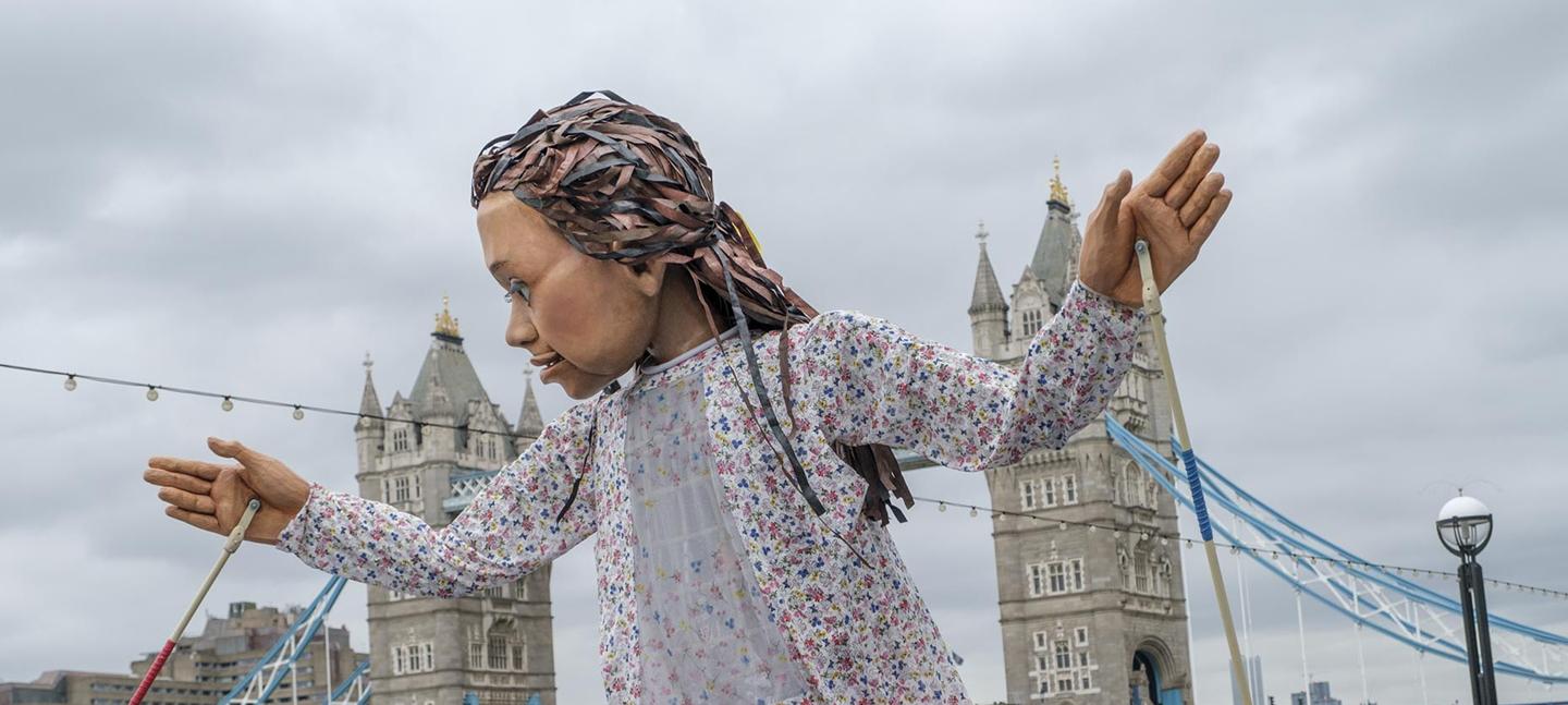 A photo of Little Amal, walking alongside the River Thames. In the background you can see Tower Bridge. The puppet looks to its right, with her hands held up and apart.