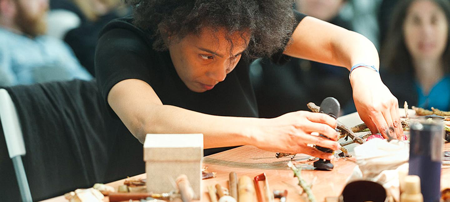 A photo of Elaine Mitchener working at a table, constructing an uknown object, In the background you can see  people sat watching.