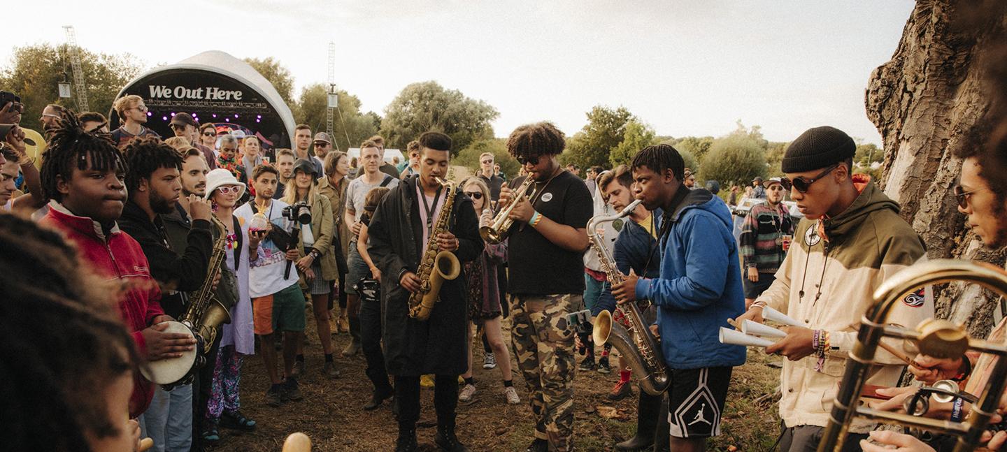 A photo of Tomorrow's Warriors performing at We Out Here Festival. A group of musicians perform on brass instruments while a crowd gathers arou.d