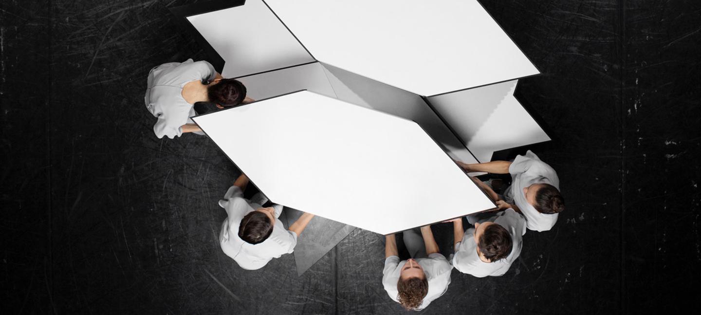 An artwork by Jasmina Cibic. An aerial view of 5 people moving a geometric, white shape across a black floor.