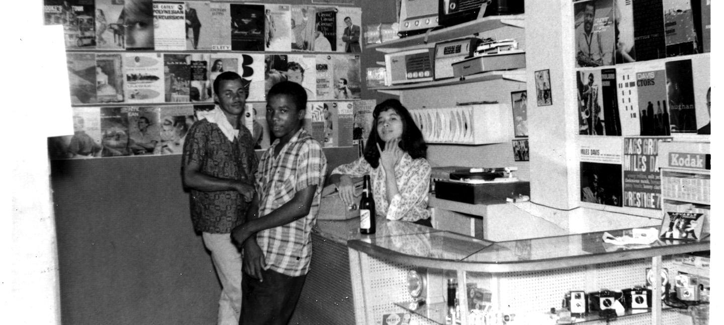 Ms Pat with customers - Randys interior 1960-61 at 36 East Street. A woman stands behind a counter in a record store, while two men stand the other side. All three people are looking at the camera.