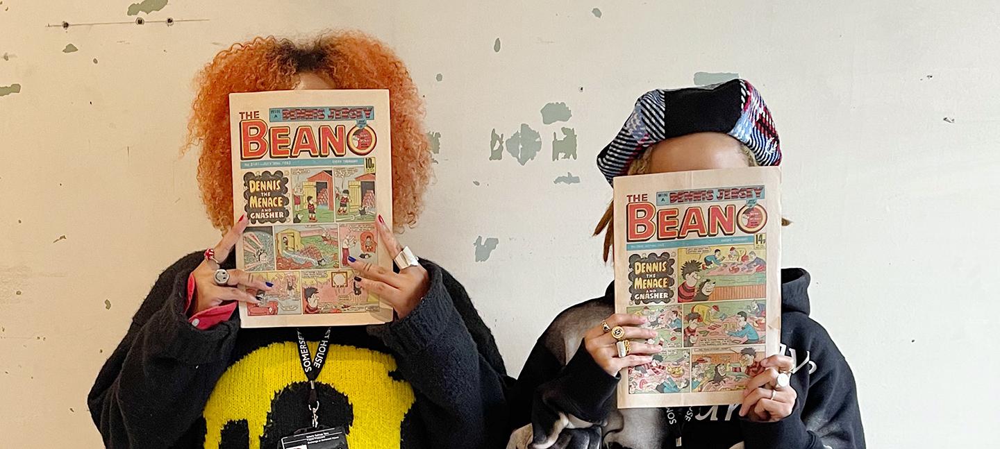 Valeria Salinas Toro and Rene Matić stand in front of the camera, holding archival prints of the Beano comic in front of their faces.