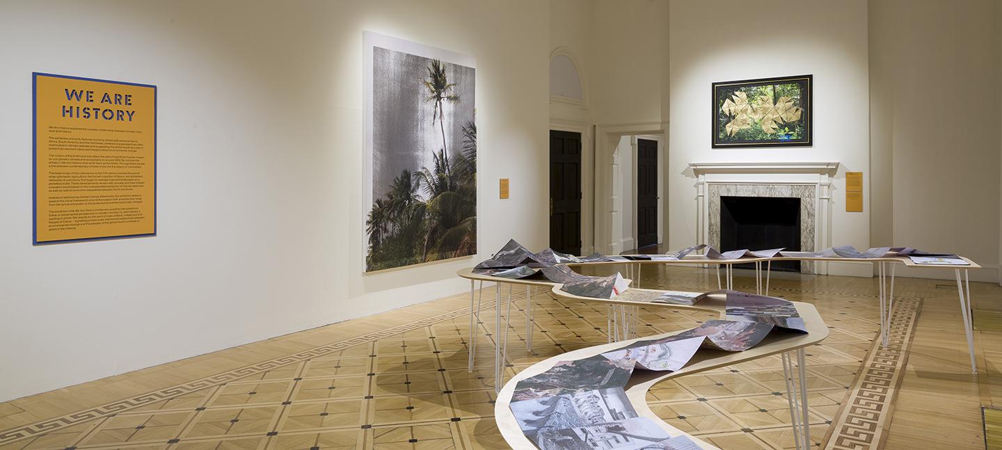 A photo of an installation view of We Are History  In the middle of the room is a work laid out on a twisting table, like the bends of a river.  On the wall are two artworks depicting tropical scenes, and an orange sign that says We Are History.