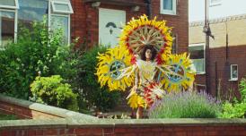 An image of a woman stood in a bright, colourful yellow, sun-like costume in a front garden.