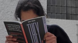 A photo of Aiysha reading a book called They by Kay Dick. The book has a black cover with a red spine, and obscured Aiysha's face.