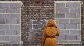 A still from 'Estate: a Reverie', 2015 by Andrea Luka Zimmerman. It shows someone in a orange coat with their hood up spraypainting a brick wall