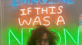 A photo of Babak Ganjei stood in front of one of his artworks. Babak stands at the front of the shot, and only his eyes and curly dark hair can be seen peering above. In the background is a neon artwork, which says 'Imagine if this were a neon'