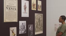 A young woman looks on at artworks carefully hung on a wall, painted a rich velvet brown.