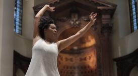 A photo of Elaine Mitchener performing. Elaine stands in a grand-looking auditorium or church, with her arms held high as she conducts music.