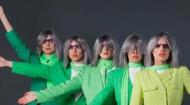 A photo of Figs in Wigs performers. Five people in grey, bob blunt cut wigs and green jackets and linen scarves around their necks stare into the camera. Each has their arm raised at a slightly different angle.