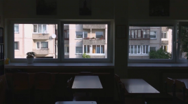 A still from Gerda Paliušytė's film Early Winter. It depicts a class room, with empty chairs and desks, with windows, looking out to buildings across the way.