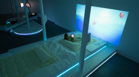 An installation image of an artwork by the collective Keiken. In a blue-lit gallery space are projections and TVs on the wall. On the floor are pillowy mattresses and cushions, surrounded by sand. People are reclining on these structures.