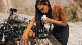 A photo of Nkisi performing on a beach at Nextones. Nkisi is shown in profile, with her decks and electronic sound equipment in front of her.