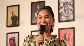 A photo of Swarzy smiling with a microphone in her hand. She stands in front of a wall of framed photos of Black creatives at an exhibition created by Swarzy, entitled Too Much Source.