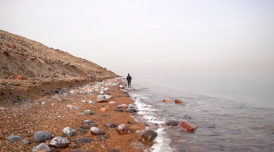 A still from a work by Dina Mimi. A small lone figure stands at the centre of shot. They are stood on a steep, pebble laden beach, with the sea on their right. It is overcast and the figure is turned away from the camera, and looks to be moving away.