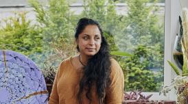 A photo of Halima Cassell. Halima is a South Asian woman, She sits in her studio, flanked by two of her works either side, smiling and looking off camera. The backdrop is full of leafy green trees.