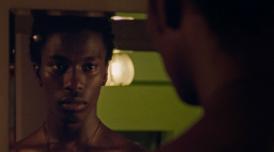 A still from a film by Iggy London. It shows a Black man, loking into a mirror, where we can see a reflection of his face.