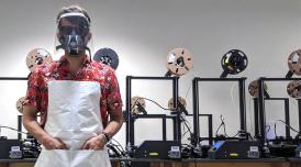 A photo of a man stood in front of 3-D printing equipment wearing a red shirt and a plastic face shield.