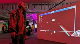 A man wearing a baseball cap, participating in a game, wears VR goggles looks at a large screen that simulates the experience of flying through space. On the right corner of the screen is a speedometer.