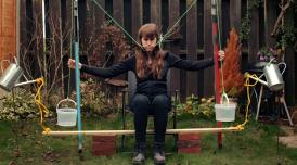 A photo of Rebecca Moss operating her smile machine. She sits in a garden, with the hooks of two coathangers hooked around the inside of her cheeks. As she pulls on ropes the coat hanger hooks pull her mouth into a smile.