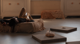 An installation of image 'We can no longer deny ourselves'. The artist lies on a fabric covered cushion with an arm above their head and knees in the air.  On the floor around them are low plinths with crystals neatly placed on them.