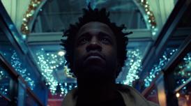 A still from the film Simeon Barclay: Defying the Norm. In it the artist Simeon Barclay, a Black man with medium length hair looks upwards as he stands in a shopping arcade in Leeds, that is lit with fairy lights in the darkness.