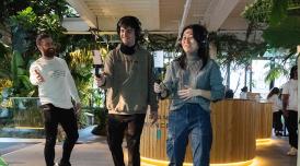 A photo of placement participants at work on 'Green Planet'. Two young people hold technical film equipment out in front of them, while someone else looks on with their arms thrown wide. There are green plants all around them.