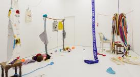 An installation view of Tyries Holder's work. Colourful scarves and fabric hang from the ceiling and there are balls of yarn on the floor.