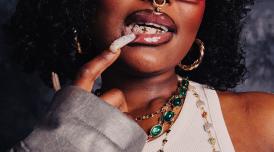 A close-up of a person who holds a manicured long acrylic nail to their lips, which are immaculately lined and lip glossed. They are wearing gold jewellery and a gold septum piercing and you can just begin to see the frames of their glasses.