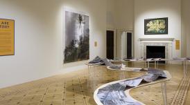 A photo of an installation view of We Are History  In the middle of the room is a work laid out on a twisting table, like the bends of a river.  On the wall are two artworks depicting tropical scenes, and an orange sign that says We Are History.