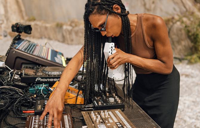 A photo of Nkisi performing on a beach at Nextones. Nkisi is shown in profile, with her decks and electronic sound equipment in front of her.