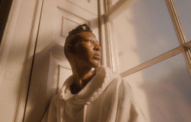 A still from Artist in Focus film of SERAFINE1369. A Black person stands sits in front of a window bathed in sunlight looking out.