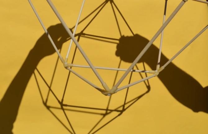 A geometric 3D shape built from interconnecting sticks is held aloft by disembodied hands of which you can see the shadows of.